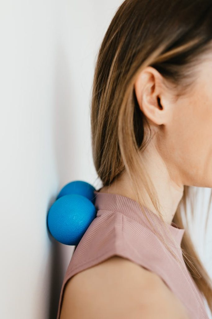 Side view of a woman using blue massage balls against a wall for neck and shoulder tension relief.